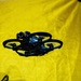 A small black FPV (First Person View) drone with ducted propeller guards is captured mid-flight against a bright yellow fabric backdrop. The drone features a forward-facing camera with an orange accent ring and enclosed rotor housings typical of indoor racing or cinematic micro drones. A partially visible graphic or logo appears in the lower-left corner of the yellow background.