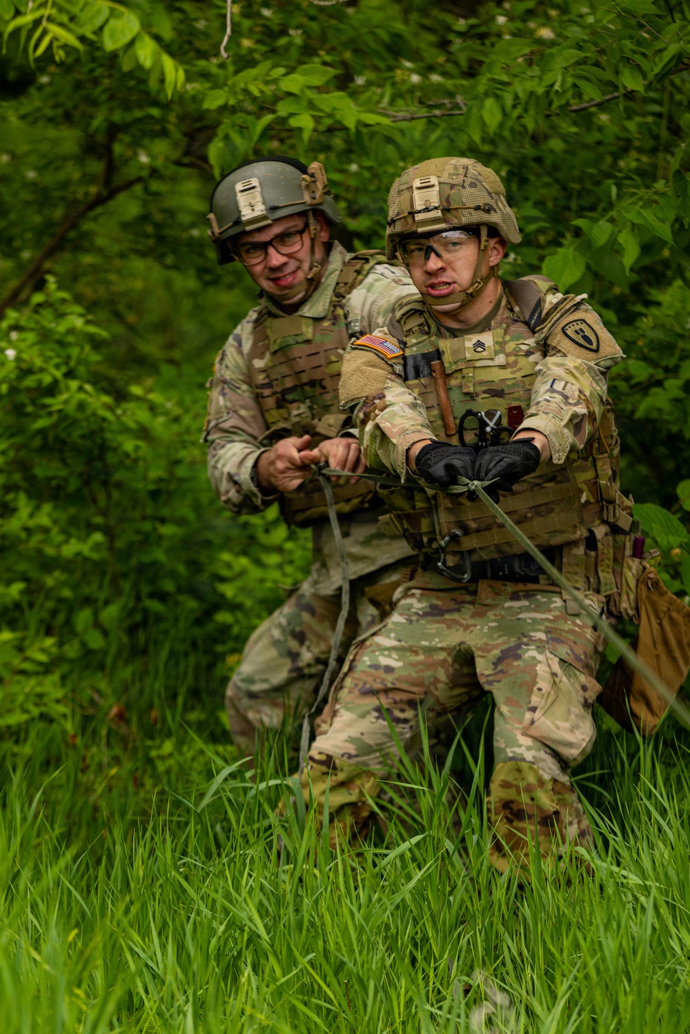 Two U.S. Army soldiers in full combat gear are pulling on a rope in a wooded, grassy outdoor environment, likely during a training exercise. Both wear Operational Camouflage Pattern (OCP) uniforms, plate carriers, and ACH helmets with accessories. The foreground soldier displays sergeant chevrons, a U.S. flag patch, and a unit shield patch on his left shoulder.