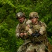 Two U.S. Army soldiers in full combat gear are pulling on a rope in a wooded, grassy outdoor environment, likely during a training exercise. Both wear Operational Camouflage Pattern (OCP) uniforms, plate carriers, and ACH helmets with accessories. The foreground soldier displays sergeant chevrons, a U.S. flag patch, and a unit shield patch on his left shoulder.