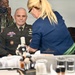 A U.S. Army officer in dress uniform is seated at a makeup/vanity station being attended to by a makeup artist with blonde hair. A second military personnel member in camouflage uniform is photographing the scene with a DSLR camera. The setting appears to be a professional makeup or media preparation area, suggesting a pre-broadcast or official portrait session.