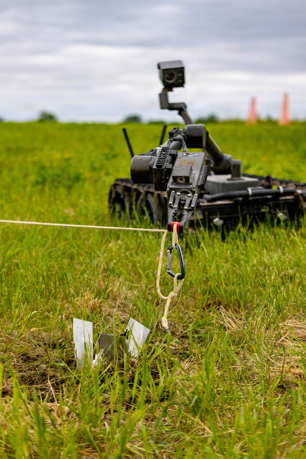 A tracked ground robot is deployed in an open grassy field, connected via a rope and carabiner to a metallic object on the ground. The robot is equipped with a camera/sensor mast and appears to be engaged in a tethered retrieval or EOD (Explosive Ordnance Disposal) type operation. Orange traffic cones are visible in the background, suggesting a controlled training or testing environment.