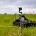 A tracked ground robot is deployed in an open grassy field, connected via a rope and carabiner to a metallic object on the ground. The robot is equipped with a camera/sensor mast and appears to be engaged in a tethered retrieval or EOD (Explosive Ordnance Disposal) type operation. Orange traffic cones are visible in the background, suggesting a controlled training or testing environment.