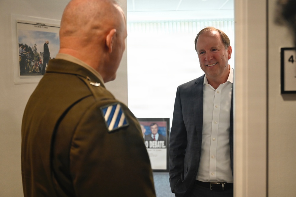 Two men are engaged in a conversation in what appears to be an office or hallway setting. One man is wearing a military uniform with a unit patch visible on the sleeve, while the other is dressed in civilian business attire (blazer and white shirt). The civilian man is smiling and appears engaged in the discussion.