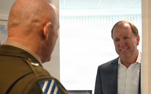 Two men are engaged in a conversation in what appears to be an office or hallway setting. One man is wearing a military uniform with a unit patch visible on the sleeve, while the other is dressed in civilian business attire (blazer and white shirt). The civilian man is smiling and appears engaged in the discussion.