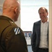 Two men are engaged in a conversation in what appears to be an office or hallway setting. One man is wearing a military uniform with a unit patch visible on the sleeve, while the other is dressed in civilian business attire (blazer and white shirt). The civilian man is smiling and appears engaged in the discussion.