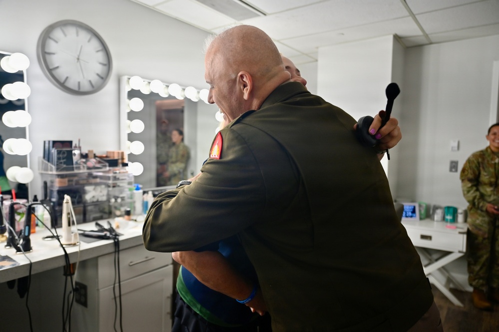 A bald individual wearing an olive/dark green military uniform is embracing another person in what appears to be a backstage makeup or dressing room. A second person in military camouflage uniform is visible in the background to the right. The setting suggests a pre- or post-event backstage environment.