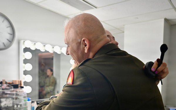 A bald individual wearing an olive/dark green military uniform is embracing another person in what appears to be a backstage makeup or dressing room. A second person in military camouflage uniform is visible in the background to the right. The setting suggests a pre- or post-event backstage environment.