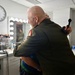 A bald individual wearing an olive/dark green military uniform is embracing another person in what appears to be a backstage makeup or dressing room. A second person in military camouflage uniform is visible in the background to the right. The setting suggests a pre- or post-event backstage environment.