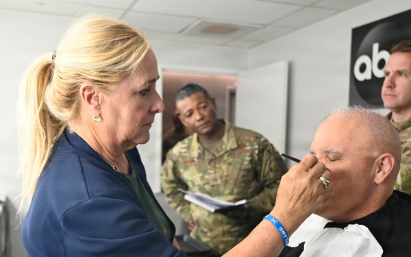 A makeup artist is applying makeup or touch-ups to a bald man seated in a styling chair, who is wearing a black cape. The setting appears to be an ABC News hair and makeup room in Washington, as indicated by the signage on the wall. Two individuals in U.S. Army camouflage uniforms are present in the background, observing the scene.