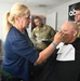 A makeup artist is applying makeup or touch-ups to a bald man seated in a styling chair, who is wearing a black cape. The setting appears to be an ABC News hair and makeup room in Washington, as indicated by the signage on the wall. Two individuals in U.S. Army camouflage uniforms are present in the background, observing the scene.