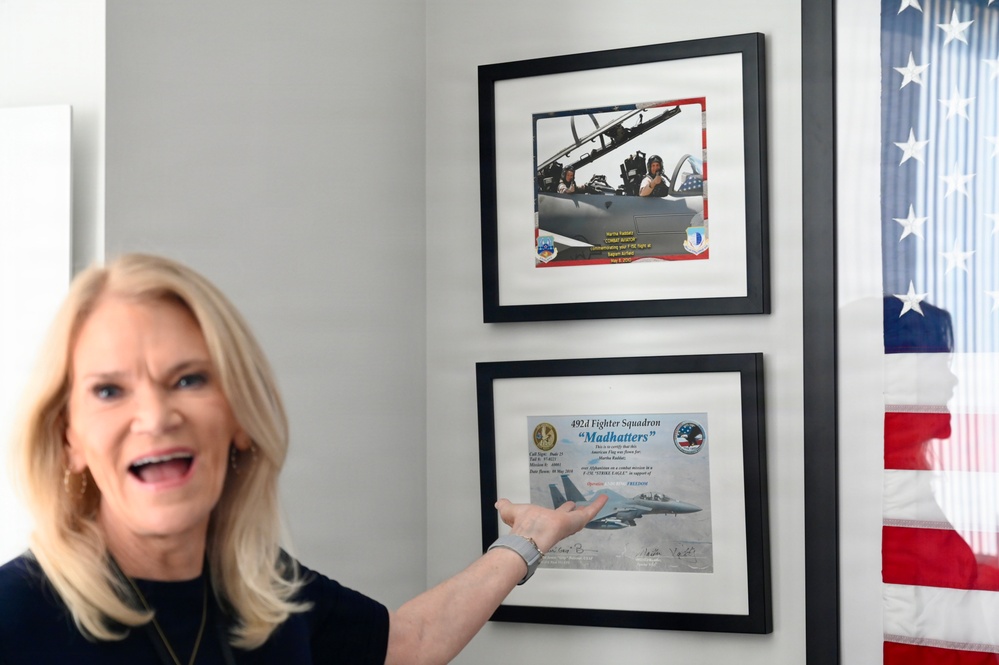 A blonde woman is enthusiastically pointing at two framed military aviation certificates/photos mounted on a wall. The frames appear to contain a photograph of someone in a fighter jet cockpit and a certificate from the 492d Fighter Squadron 'Madhatters'. An American flag is partially visible on the right side of the image.