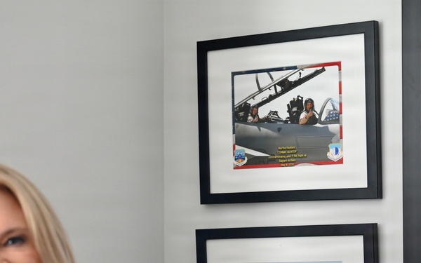 A blonde woman is enthusiastically pointing at two framed military aviation certificates/photos mounted on a wall. The frames appear to contain a photograph of someone in a fighter jet cockpit and a certificate from the 492d Fighter Squadron 'Madhatters'. An American flag is partially visible on the right side of the image.
