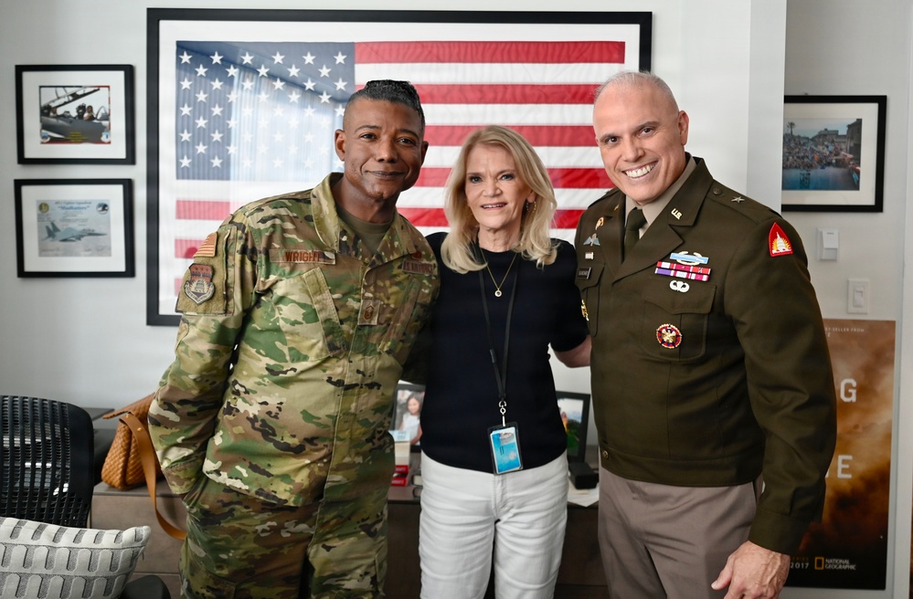Three individuals pose for a photo in front of a large framed American flag. Two are in U.S. military uniforms — one in Army Combat Uniform (ACU/OCP camouflage) and one in Army Service Uniform (green dress uniform) — flanking a civilian woman in casual attire. The setting appears to be an office or media environment, with framed photographs and a National Geographic poster visible in the background.