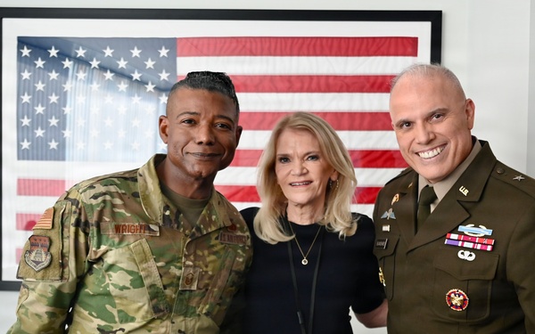 Three individuals pose for a photo in front of a large framed American flag. Two are in U.S. military uniforms — one in Army Combat Uniform (ACU/OCP camouflage) and one in Army Service Uniform (green dress uniform) — flanking a civilian woman in casual attire. The setting appears to be an office or media environment, with framed photographs and a National Geographic poster visible in the background.