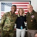 Three individuals pose for a photo in front of a large framed American flag. Two are in U.S. military uniforms — one in Army Combat Uniform (ACU/OCP camouflage) and one in Army Service Uniform (green dress uniform) — flanking a civilian woman in casual attire. The setting appears to be an office or media environment, with framed photographs and a National Geographic poster visible in the background.