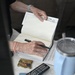 An elderly person's hands are shown opening a hardcover book, apparently preparing to sign or inscribe it. On the table nearby are a gold badge/coin, a TV remote control, a light blue tumbler with a straw, and some papers. The setting appears to be a casual indoor environment.