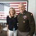 Two individuals pose together in what appears to be an office setting. One person is wearing a U.S. Army dress uniform with multiple medals, badges, and insignia, while the other is a civilian woman wearing a navy top and white pants with a lanyard/badge. A large framed American flag is prominently displayed on the wall behind them.