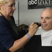 A makeup artist is applying makeup to a man seated in a makeup chair, preparing him for an on-camera appearance. The setting is a professional makeup studio affiliated with ABC News in Washington, DC. The man is wearing a protective cape and appears calm while the artist works on his face with a brush.