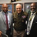 Three men pose together for a photo in what appears to be a military office setting. The center individual is wearing a U.S. Army dress uniform with multiple medals, badges, and a name tag reading 'BLANCHARD'. The two men flanking him are dressed in civilian business attire.