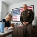 A woman with blonde hair is seated at a desk signing what appears to be a book, while a U.S. Army officer in dress green uniform stands beside her smiling. The setting appears to be an office with an American flag artwork on the wall and various framed photos on a credenza. A poster partially visible on the right reads 'The Long Road Ho[me]', suggesting a possible book signing or media-related event.