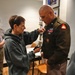A woman in a gray hoodie is attaching a lavalier microphone to the uniform of a U.S. Army officer wearing an olive green dress uniform with multiple medals, ribbons, and rank insignia. The setting appears to be an indoor office or media room, suggesting the officer is being prepared for a television or media interview. A second person wearing a blue lanyard badge is visible in the background.