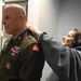 A U.S. Army officer in dress uniform is having insignia or rank devices pinned onto his shoulder/collar area by two civilians. The officer wears a green Class A uniform with multiple medals, ribbons, a unit patch, and what appears to be a general officer star visible on his collar. This appears to be a military promotion or award ceremony taking place in an indoor office or library setting.