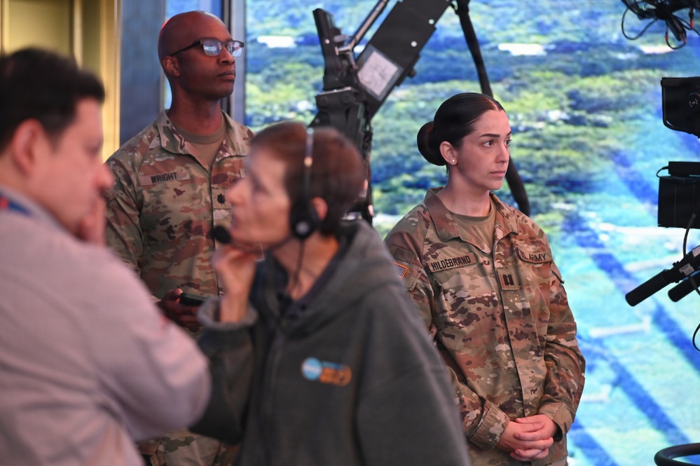Two U.S. Army soldiers in OCP (Operational Camouflage Pattern) uniforms stand in what appears to be a broadcast or media studio environment. The soldier on the right has the name tape 'HILDEBRAND' and a captain's rank insignia, while the soldier in the background bears the name tape 'WRIGHT'. They appear to be observing a broadcast or media production in progress.