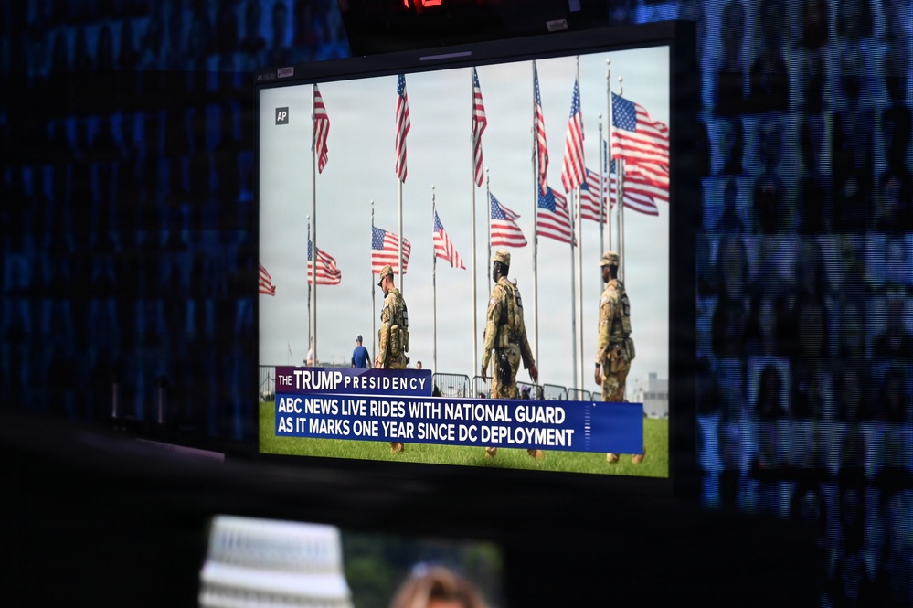 A television monitor displays an ABC News broadcast under the banner 'The Trump Presidency,' showing National Guard soldiers standing among multiple American flags. The chyron reads 'ABC News Live Rides With National Guard As It Marks One Year Since DC Deployment.' The monitor appears to be in a darkened studio or control room environment with blue-lit screens visible in the background.