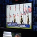 A television monitor displays an ABC News broadcast under the banner 'The Trump Presidency,' showing National Guard soldiers standing among multiple American flags. The chyron reads 'ABC News Live Rides With National Guard As It Marks One Year Since DC Deployment.' The monitor appears to be in a darkened studio or control room environment with blue-lit screens visible in the background.