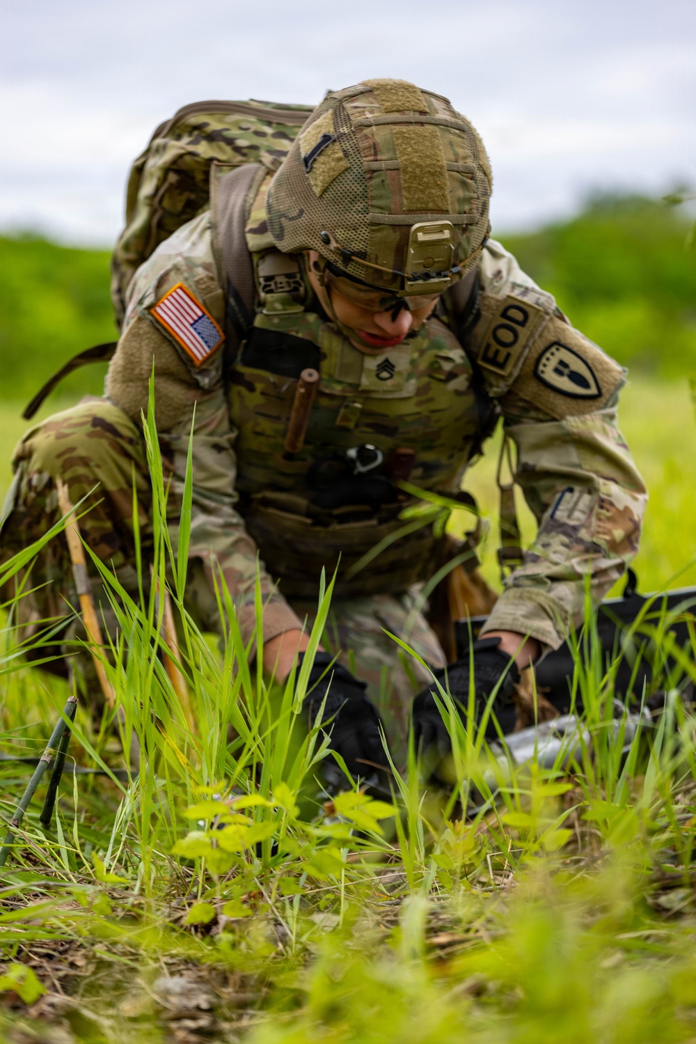 A U.S. Army EOD (Explosive Ordnance Disposal) soldier, identifiable by rank insignia of Staff Sergeant, is kneeling in a grassy field while working on equipment or a device on the ground. The soldier is wearing full MultiCam combat uniform, tactical helmet with NVG mount, body armor, and a large rucksack. The setting appears to be an outdoor training or operational environment.