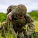 A U.S. Army EOD (Explosive Ordnance Disposal) soldier, identifiable by rank insignia of Staff Sergeant, is kneeling in a grassy field while working on equipment or a device on the ground. The soldier is wearing full MultiCam combat uniform, tactical helmet with NVG mount, body armor, and a large rucksack. The setting appears to be an outdoor training or operational environment.