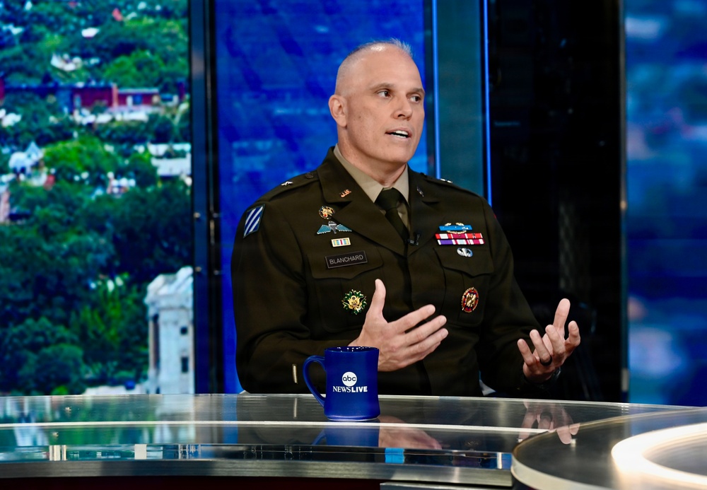 A U.S. Army officer named Blanchard, as indicated by the name tape on his uniform, is seated at a news desk during a live broadcast on ABC News Live. He is wearing an olive green Army service uniform adorned with multiple medals, badges, and insignia, and appears to be speaking or being interviewed. The setting is a modern television studio with a blue-lit backdrop and a city/landscape view visible through a window.