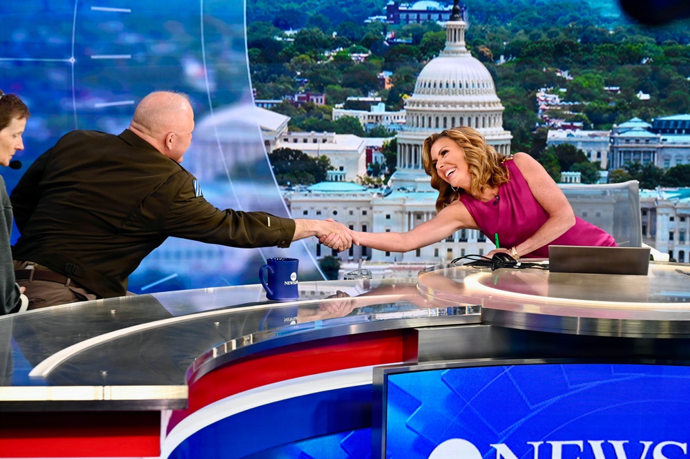 A television news broadcast set featuring two individuals shaking hands across an anchor desk. A woman in a pink/magenta sleeveless dress leans forward smiling while shaking hands with a bald man in a dark olive military-style shirt. The backdrop displays a large screen image of the U.S. Capitol building in Washington, D.C.