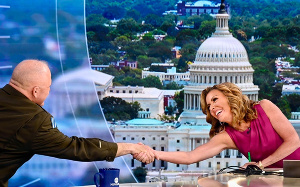 A television news broadcast set featuring two individuals shaking hands across an anchor desk. A woman in a pink/magenta sleeveless dress leans forward smiling while shaking hands with a bald man in a dark olive military-style shirt. The backdrop displays a large screen image of the U.S. Capitol building in Washington, D.C.