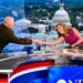 A television news broadcast set featuring two individuals shaking hands across an anchor desk. A woman in a pink/magenta sleeveless dress leans forward smiling while shaking hands with a bald man in a dark olive military-style shirt. The backdrop displays a large screen image of the U.S. Capitol building in Washington, D.C.