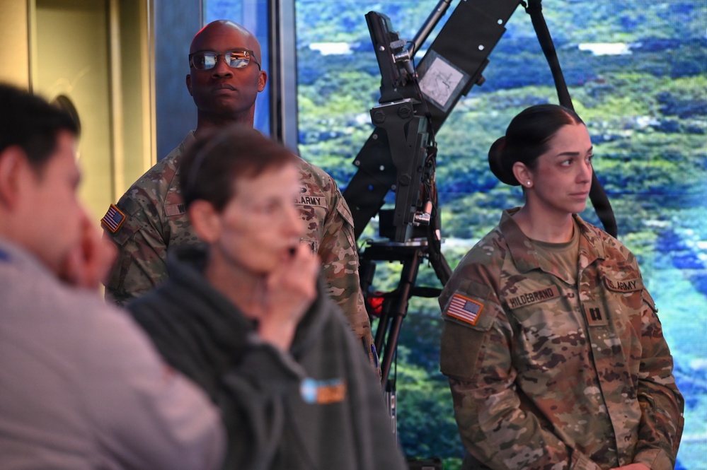 Two U.S. Army soldiers in OCP (Operational Camouflage Pattern) uniforms stand attentively in what appears to be an indoor briefing or presentation setting. A female soldier with the name tape 'HILDEBRAND' and a captain's rank insignia is visible on the right, while a male soldier stands behind a mounted equipment piece in the center. Several civilians are also present in the foreground.