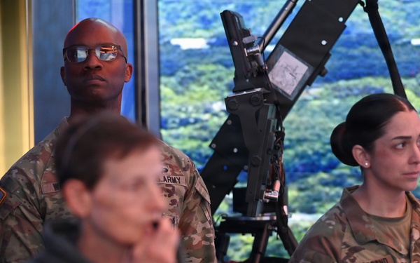 Two U.S. Army soldiers in OCP (Operational Camouflage Pattern) uniforms stand attentively in what appears to be an indoor briefing or presentation setting. A female soldier with the name tape 'HILDEBRAND' and a captain's rank insignia is visible on the right, while a male soldier stands behind a mounted equipment piece in the center. Several civilians are also present in the foreground.