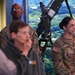 Two U.S. Army soldiers in OCP (Operational Camouflage Pattern) uniforms stand attentively in what appears to be an indoor briefing or presentation setting. A female soldier with the name tape 'HILDEBRAND' and a captain's rank insignia is visible on the right, while a male soldier stands behind a mounted equipment piece in the center. Several civilians are also present in the foreground.