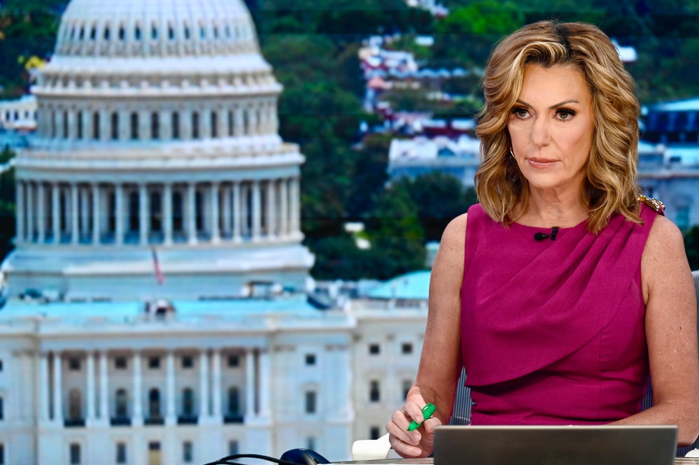 A woman is seated at what appears to be a news broadcast desk, wearing a magenta/fuchsia sleeveless dress with a lapel microphone. She is positioned in front of a backdrop or window showing the United States Capitol building in Washington D.C. She holds a green marker and appears to be in a professional broadcast setting.