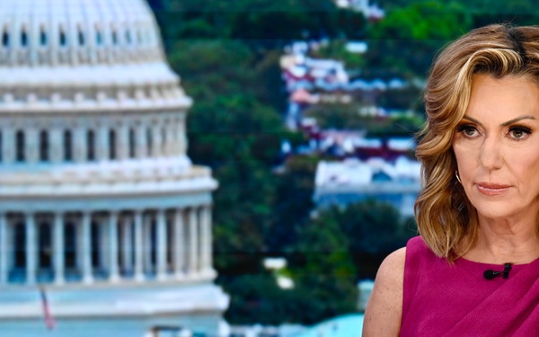 A woman is seated at what appears to be a news broadcast desk, wearing a magenta/fuchsia sleeveless dress with a lapel microphone. She is positioned in front of a backdrop or window showing the United States Capitol building in Washington D.C. She holds a green marker and appears to be in a professional broadcast setting.