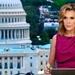 A woman is seated at what appears to be a news broadcast desk, wearing a magenta/fuchsia sleeveless dress with a lapel microphone. She is positioned in front of a backdrop or window showing the United States Capitol building in Washington D.C. She holds a green marker and appears to be in a professional broadcast setting.
