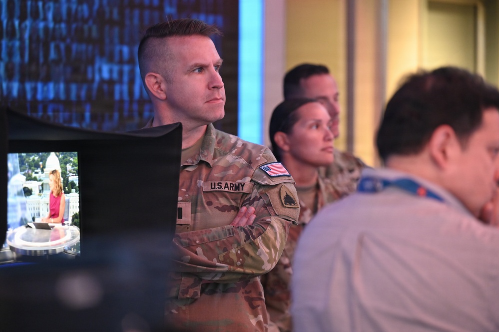 The image shows U.S. Army personnel in OCP (Operational Camouflage Pattern) uniforms standing in what appears to be a media or operations center environment. At least two soldiers are visible in the foreground, with a civilian also present to the right. A television monitor in the background appears to be displaying a news broadcast featuring the U.S. Capitol building.