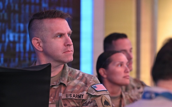 The image shows U.S. Army personnel in OCP (Operational Camouflage Pattern) uniforms standing in what appears to be a media or operations center environment. At least two soldiers are visible in the foreground, with a civilian also present to the right. A television monitor in the background appears to be displaying a news broadcast featuring the U.S. Capitol building.