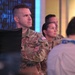 The image shows U.S. Army personnel in OCP (Operational Camouflage Pattern) uniforms standing in what appears to be a media or operations center environment. At least two soldiers are visible in the foreground, with a civilian also present to the right. A television monitor in the background appears to be displaying a news broadcast featuring the U.S. Capitol building.