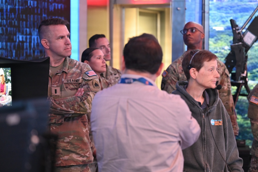 A group of U.S. Army personnel in OCP (Operational Camouflage Pattern) uniforms are visiting what appears to be a television broadcast studio or news control room. They are being guided or briefed by a civilian staff member wearing a headset and a hoodie with visible text. The setting includes broadcast equipment, monitors, and a large scenic backdrop screen.