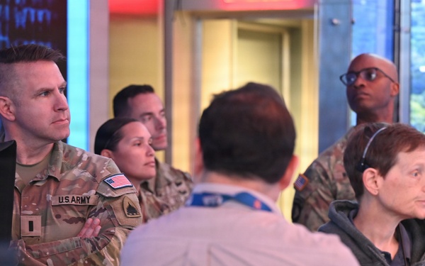 A group of U.S. Army personnel in OCP (Operational Camouflage Pattern) uniforms are visiting what appears to be a television broadcast studio or news control room. They are being guided or briefed by a civilian staff member wearing a headset and a hoodie with visible text. The setting includes broadcast equipment, monitors, and a large scenic backdrop screen.