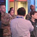 A group of U.S. Army personnel in OCP (Operational Camouflage Pattern) uniforms are visiting what appears to be a television broadcast studio or news control room. They are being guided or briefed by a civilian staff member wearing a headset and a hoodie with visible text. The setting includes broadcast equipment, monitors, and a large scenic backdrop screen.