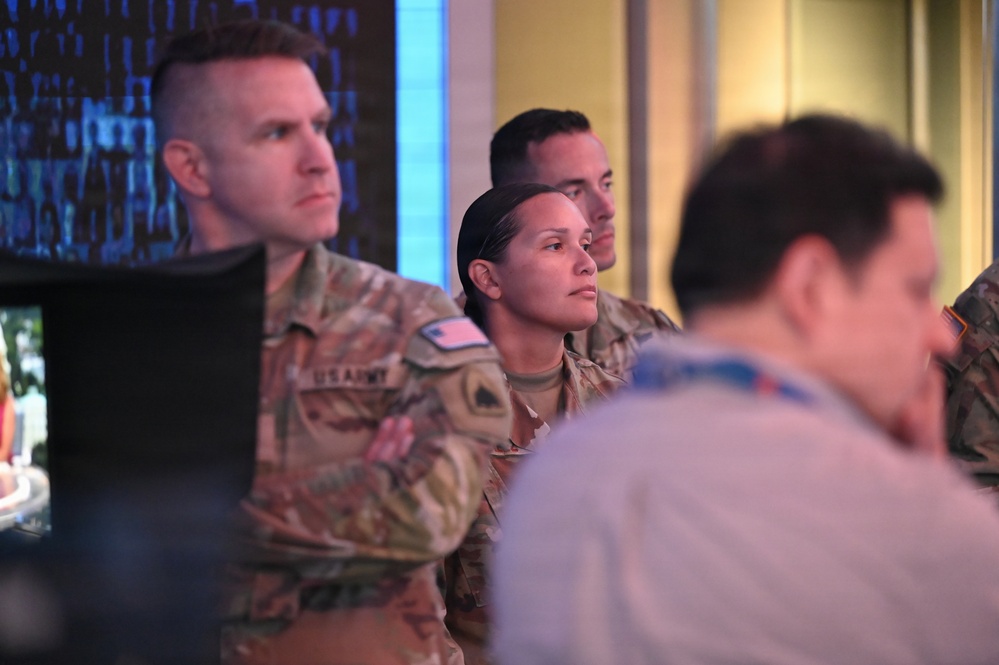 The image shows multiple U.S. Army personnel in OCP (Operational Camouflage Pattern) uniforms standing and observing something off-camera, likely in a briefing or operations center environment. A digital display with blue data/code patterns is visible in the background. The personnel appear attentive and focused.