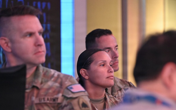 The image shows multiple U.S. Army personnel in OCP (Operational Camouflage Pattern) uniforms standing and observing something off-camera, likely in a briefing or operations center environment. A digital display with blue data/code patterns is visible in the background. The personnel appear attentive and focused.