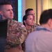 The image shows multiple U.S. Army personnel in OCP (Operational Camouflage Pattern) uniforms standing and observing something off-camera, likely in a briefing or operations center environment. A digital display with blue data/code patterns is visible in the background. The personnel appear attentive and focused.